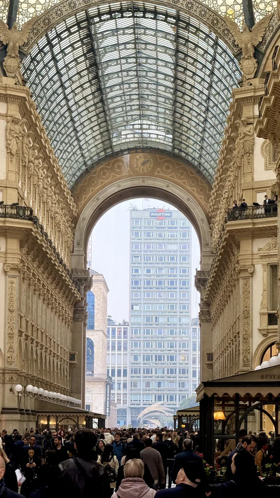 Crowd under the glass vault of the Galleria Vittorio Emanuele II in Milan, with the Martini tower framed by the arch.