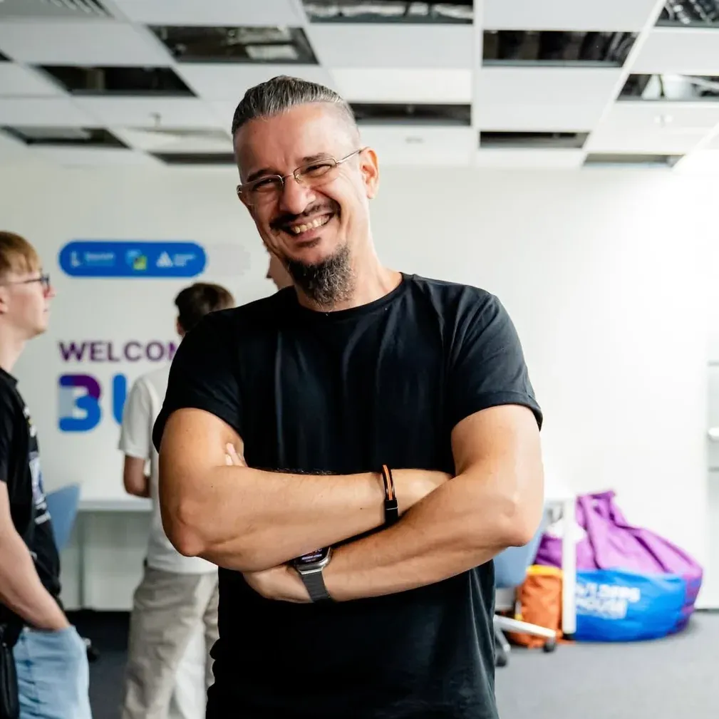Sebastian Cochinescu smiling with his arms crossed at How to Web Demo Nights 2026, in a bright office with people talking behind him.