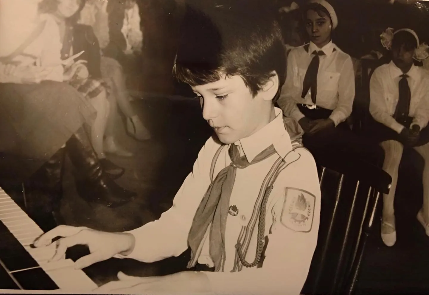 Black-and-white photo of a young boy in a pioneer uniform playing piano while other children watch.