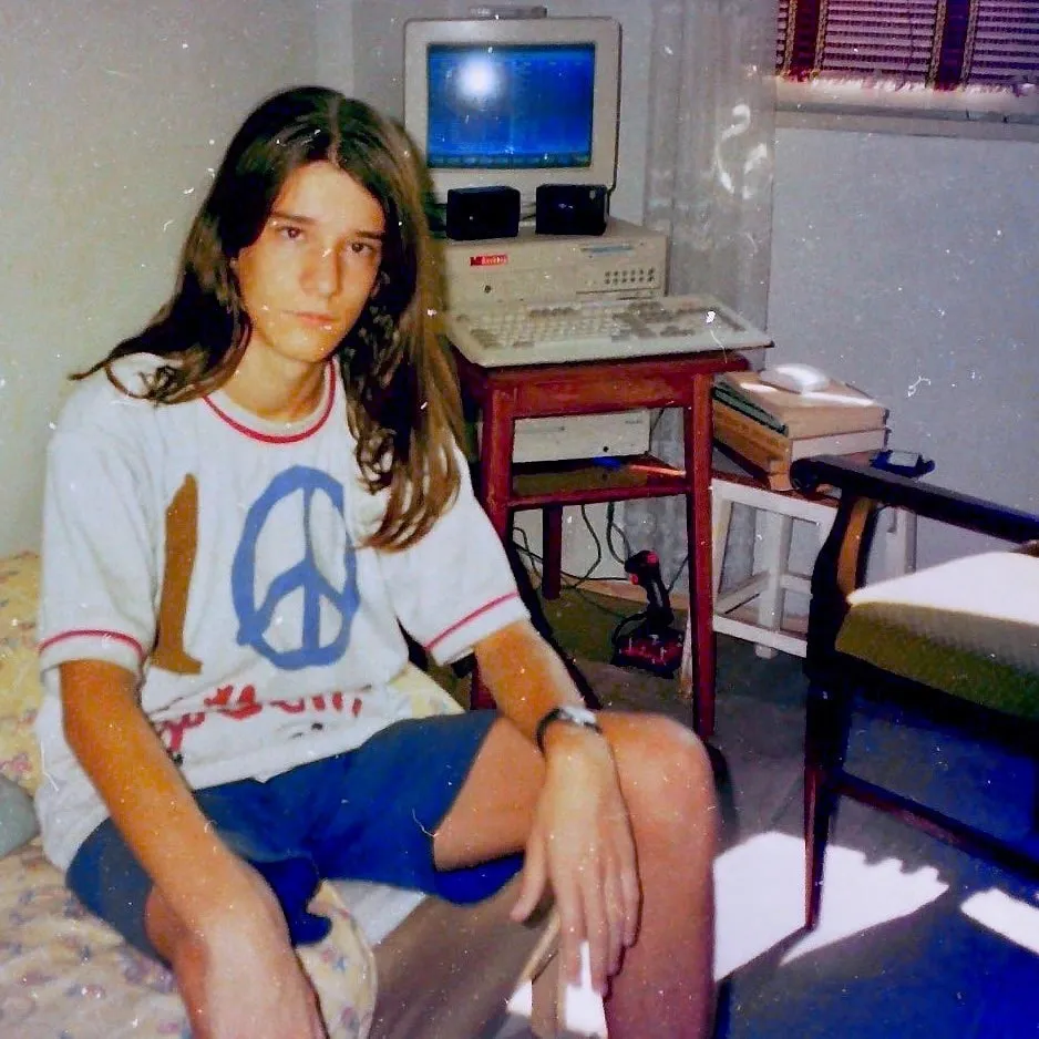 Vintage photo of a long-haired teenager sitting beside an old CRT computer on a wooden desk.