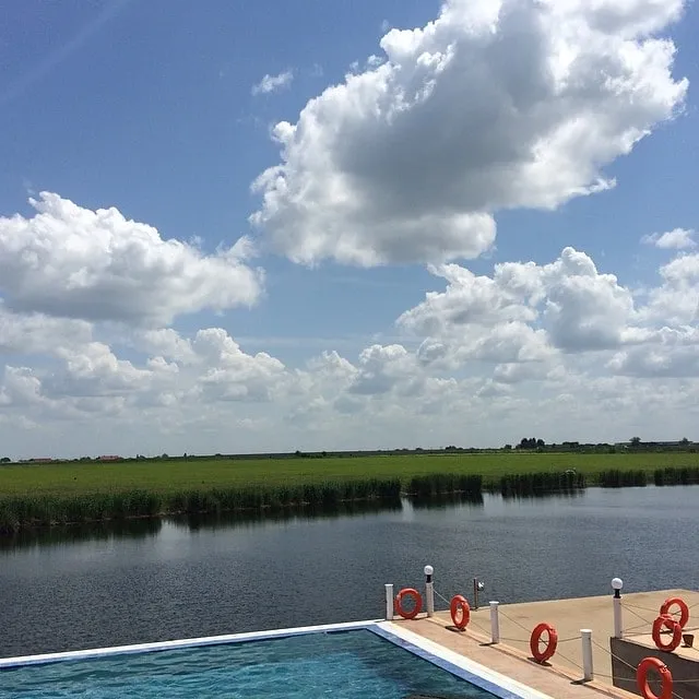 Swimming pool with orange life rings, overlooking a lake and green fields beneath a blue sky with white clouds.