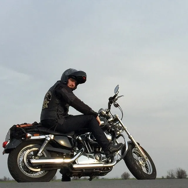 A man in a black helmet and leather vest sits on a motorcycle against a pale sky.